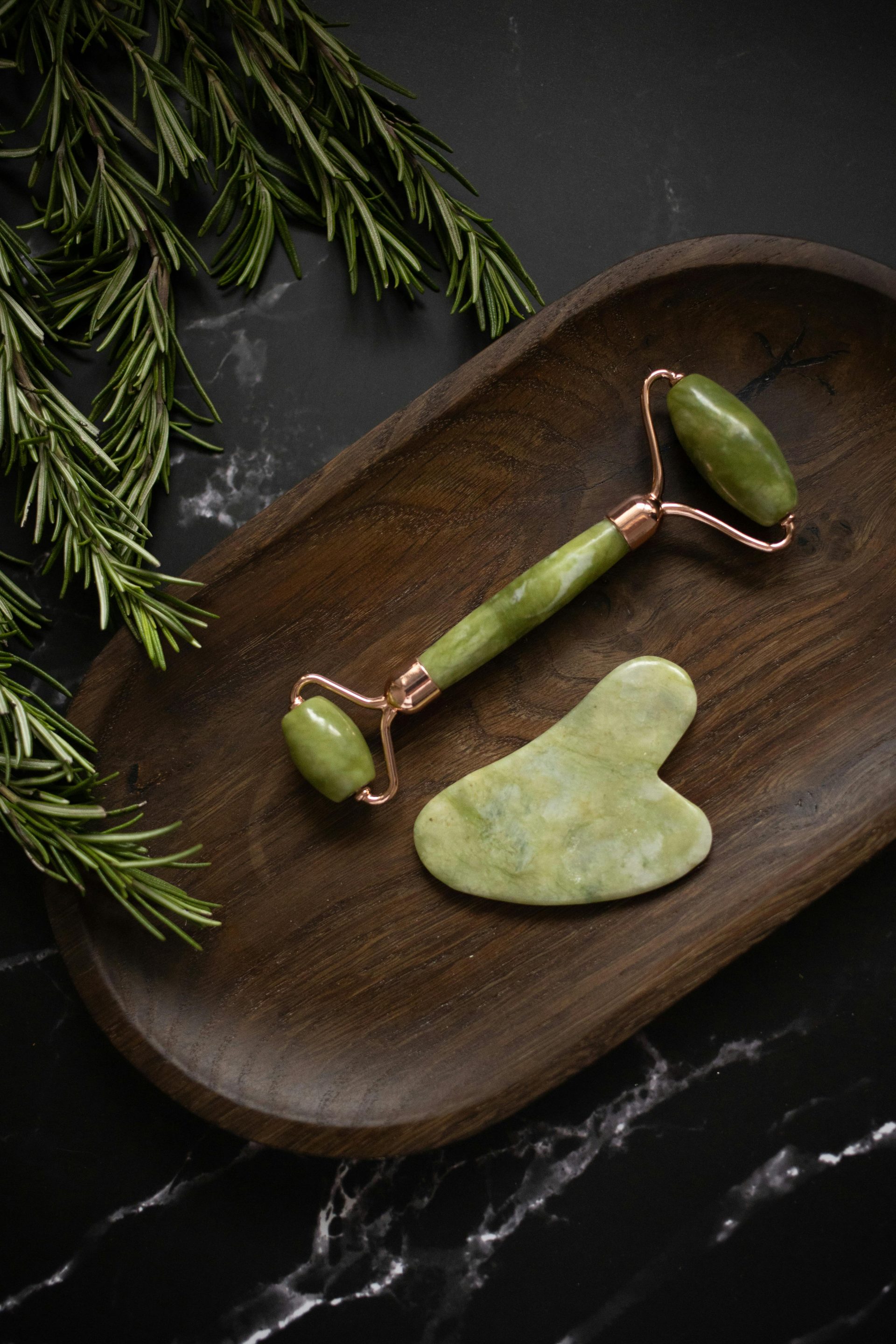 Vue de dessus d'un rouleau de jade et d'un outil de massage Gua Sha placés sur une plaque de bois posée sur une table en marbre noir avec des brins de romarin frais